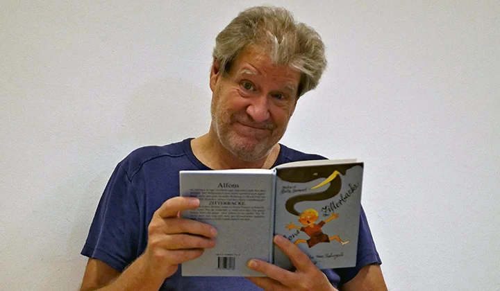 Schauspieler Marcus Staiger schaut verschmitzt in die Kamera und hält das Buch Alfons Zitterbacke von Gerhard Holtz-Baumert dabei in den Händen
