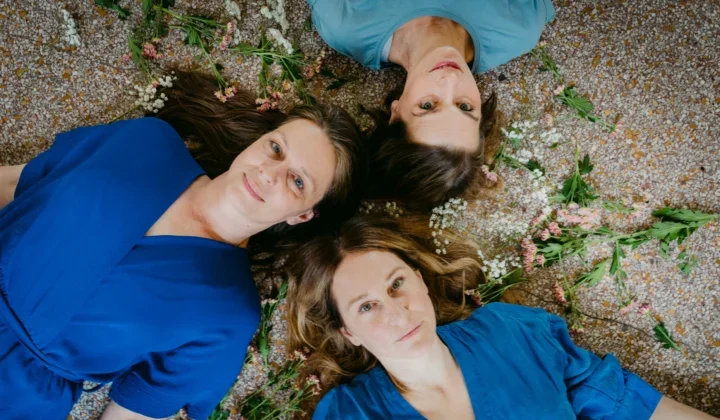 Drei Frauen in blauen Kleidern liegen sternförmig auf dem Rücken auf einem feinen Kiesboden, der mit Blumen bestreut, und blicken in den Himmel.