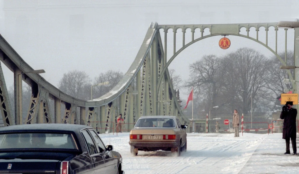 Die Glienicker Brücke im Winter. Auf der Brücke sind Sperren. Zwei Limousinen sind darauf zu sehen und 3 Personen. Eine Person fotografiert die Wagen.